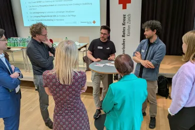 Rund um einen Stehtisch stehen mehrere Personen und hören gespannt zu. Es wird über ein Programm vom Jugendrotkreuz Zürich erzählt. 