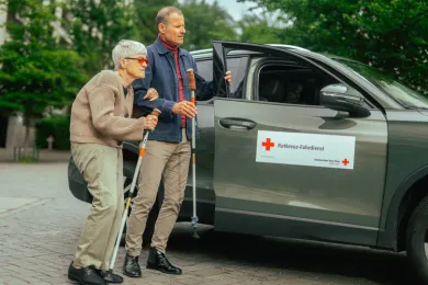 Ein Fahrdienst-Freiwilliger unterstützt eine Seniorin auf dem Weg zum Auto.