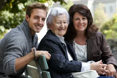 Zwei jüngere Personen sitzen mit einer Seniorin auf einer Bank und lachen zusammen.