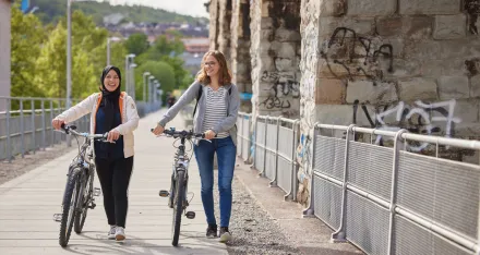 Zwei weibliche Personen spazieren zusammen. Sie stossen dabei ihre Velos.