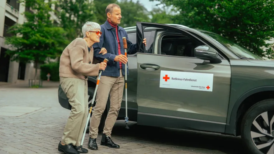 Ein Fahrdienst-Freiwilliger unterstützt eine Seniorin auf dem Weg zum Auto.
